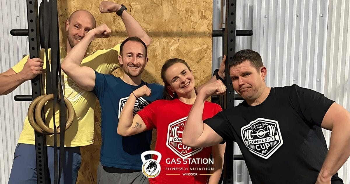 Four fitness coaches flexing their arms and smiling in a gym setting, standing in front of workout equipment with wooden panels and metal walls. Three wear community-branded workout shirts while one wears yellow.